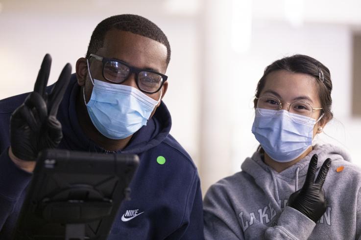 A man and woman side by side wearing disposable masks and black latex gloves, flashing the peace sign