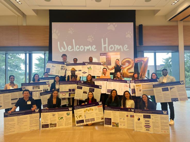 A group of 23 DGH MPH students holding their project posters stand in front of a projector screen that says Welcome Home