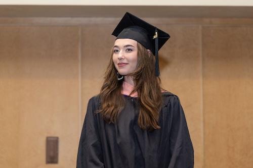 Outstanding Master's Student Brekken Cogswell wears black graduation attire and stands in front of the auditorium.