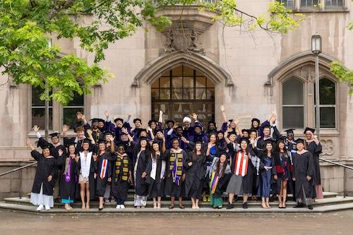 All DGH graduates wear graduation regalia and throw their hands in the air for a group shot in front of an ornate UW building.