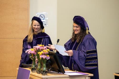 Both wearing purple graduation regalia, Professor Alison Drake watches Kyle Daniels, graduate of the Doctor of Global Health Leadership and Practice Program, give her speech behind the podium.