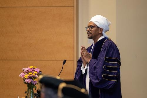 Mohamed Tawfig Albirair, Implementation Science PhD graduate, wears purple graduation regalia and stands behind the podium while delivering his speech.