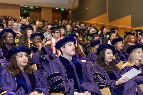 A group of PhD graduate students wearing purple graduation attire sit in the auditorium and listen to the speaker.